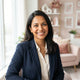 A friendly South Asian parent smiling for a portrait in a bright, modern home setting with soft rose and pink decor.