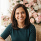 A friendly South Asian mother smiling in a bright, modern setting with soft pink and white accents.