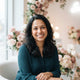 A friendly South Asian mother smiling in a bright, modern room with soft rose and blush pink decor.