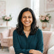 A smiling South Asian mother in a brightly lit, modern home setting with soft pink and white decor.